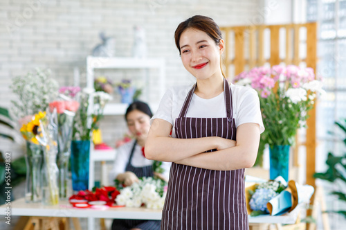 Portrait studio shot of Millennial Asian young female flower shopkeeper decorator florist employee worker in apron standing smiling in front blurred workshop table in floral store.