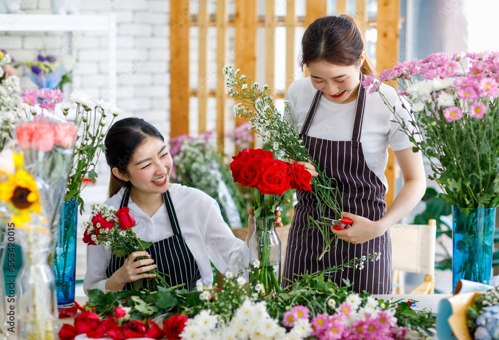 Millennial Asian young female flower shopkeeper decorator florist worker in apron smiling holding flower helping bunch bouquet colleague arranging decorating stalk in floral store.