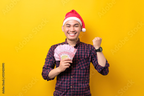 Excited young Asian man in Santa hat holding fan of cash money and raising fist, celebrating success on yellow studio background. Happy New Year 2023 celebration merry holiday concept