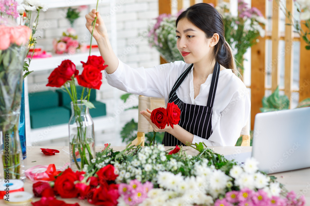 Millennial Asian young female flower shopkeeper decorator florist employee worker in apron sitting smiling at workshop table arranging decorating red roses flowers into glass vase in floral store