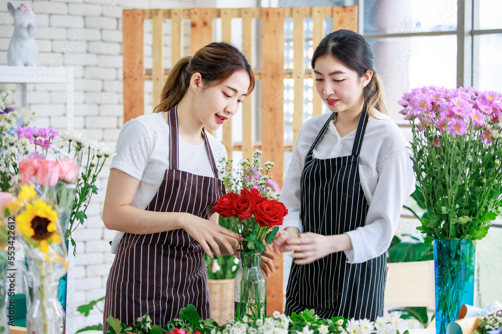 Millennial Asian young female flower shopkeeper decorator florist worker in apron smiling holding flower bunch bouquet colleague arranging decorating stalk in floral store.
