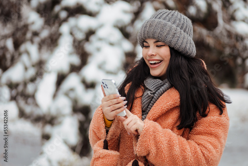 Pretty young long haired woman with amazing smile using her mobile phone at a snowy winter park