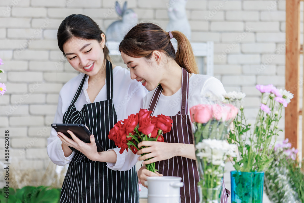 Millennial Asian young professional flower shopkeeper florist employee worker wearing apron standing smiling holding red roses bunch bouquet while colleague using tablet computer in floral store