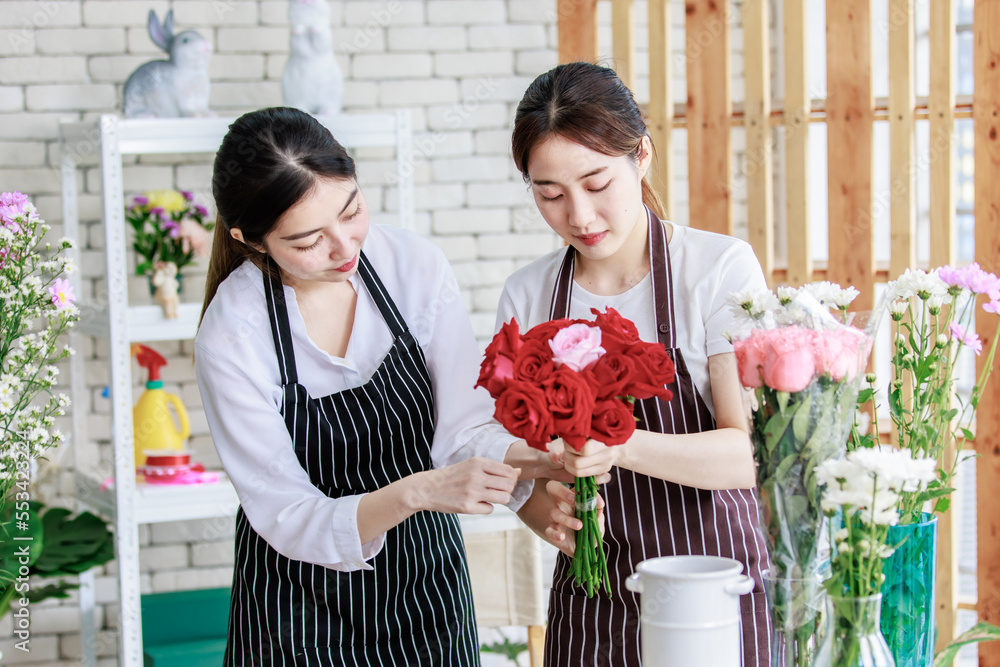 Millennial Asian young professional female florist shopkeeper employee worker wearing apron holding red roses bunch bouquet while colleague using ribbon wrapping flowers stalk together in floral shop