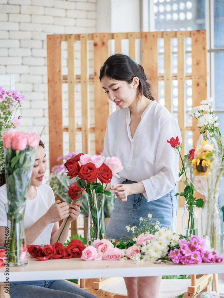 Millennial Asian young beautiful professional female flower shopkeeper decorator florist worker smiling holding flower bunch bouquet colleague arranging decorating stalk in floral store.