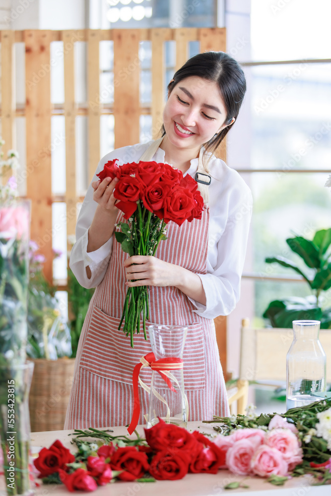 Millennial Asian young professional flower shopkeeper florist employee worker wearing apron standing smiling closed eye holding sending red roses bunch bouquet posing taking photo in floral store