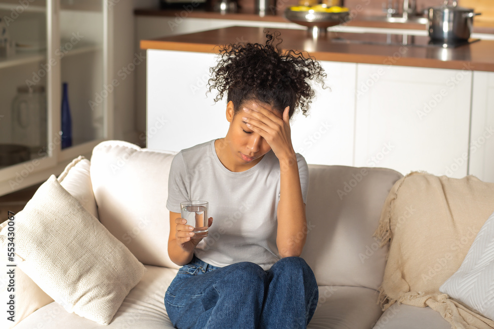 Unhappy African American woman sitting on the couch with a glass of water feeling bad, experiencing headache and dizziness due to illness Unhappy African American woman sitting on the couch with a glass of water feeling bad, experiencing headache and dizziness due to illness