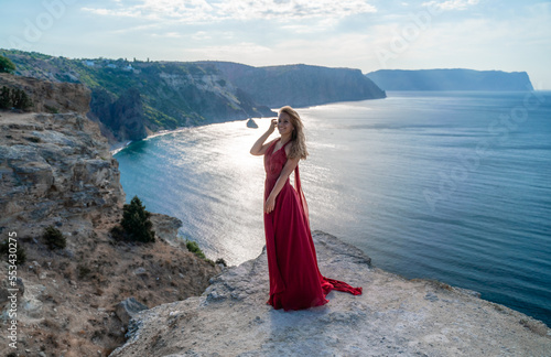 A girl with flowing hair in a long red dress stands on a rock above the sea. The stone can be seen in the sea. Sunny path to the sea from the sun.