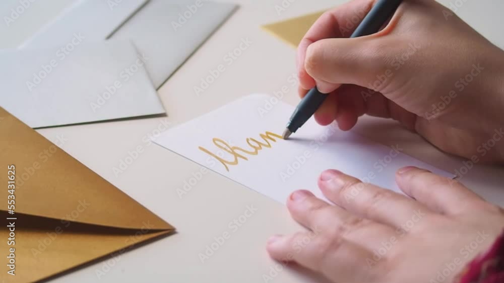 Close up of hands of woman writing a thank you note on a card, in golden calligraphy, expressing gratitude for a gift or service, with fancy golden and silver envelopes on her desk