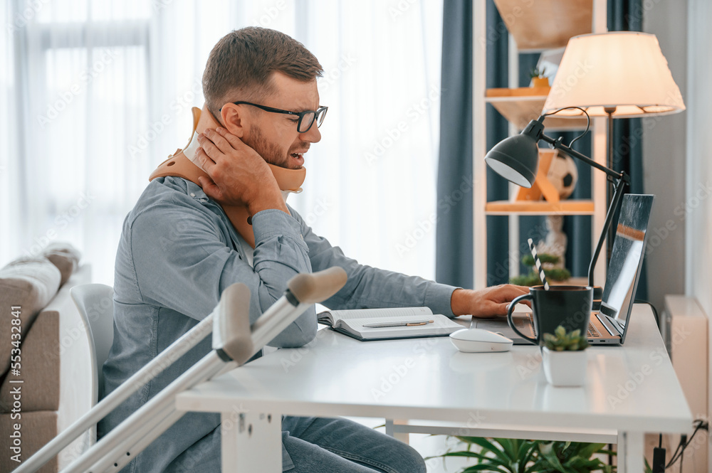 Neck pain. Sitting by table with laptop. Man with crutches is at home indoors. Having injuries