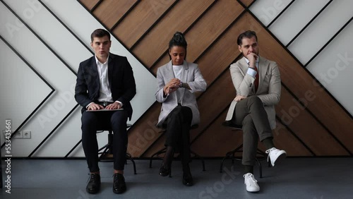 anxious multi-sex multinational job seekers waiting for an invitation for an interview in a sterile office sitting on chairs in the hallway
