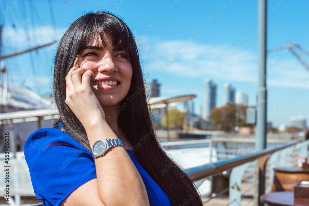 Obraz premium happy young smiling latin woman talking on the phone, sitting outside a restaurant.