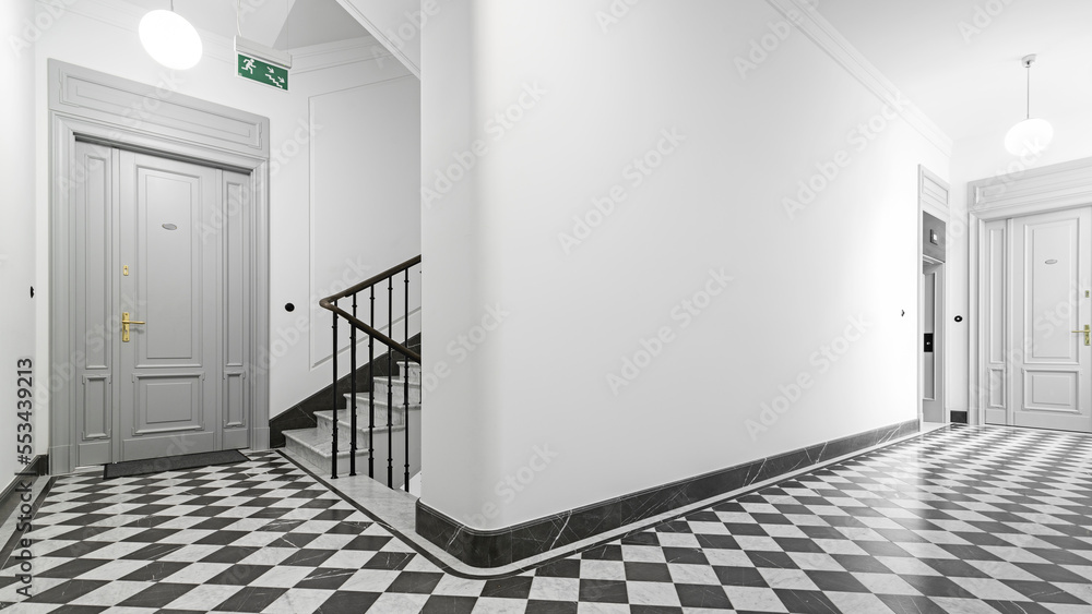 Fototapeta premium Klatka schodowa w stylu klasycznym. Stalowa balustrada z drewnianymi elementami. Duże szare drzwi wejściowe do mieszkania. Przestrony korytarz z windą