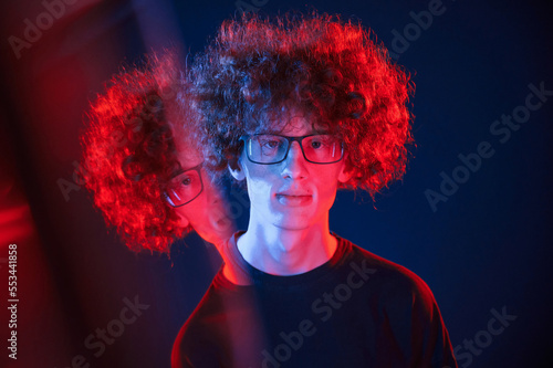 Glass reflection on the left side. Young man with curly hair is indoors illuminated by neon lighting