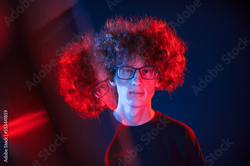 Glass reflection on the left side. Young man with curly hair is indoors illuminated by neon lighting