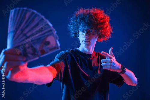Holding money. Young man with curly hair is indoors illuminated by neon lighting