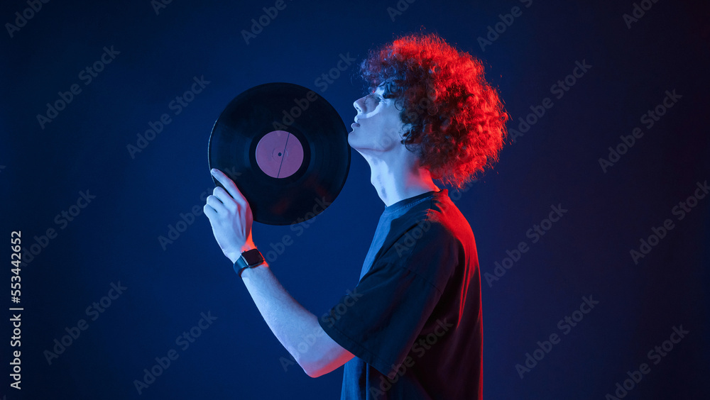 Musician holding vinyl. Young man with curly hair is indoors illuminated by neon lighting Musician holding vinyl. Young man with curly hair is indoors illuminated by neon lighting