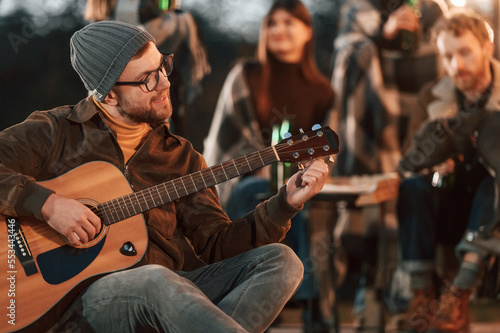 Man in hat is sitting and playing guitar. Group of people is spending time together on the backyard at evening time