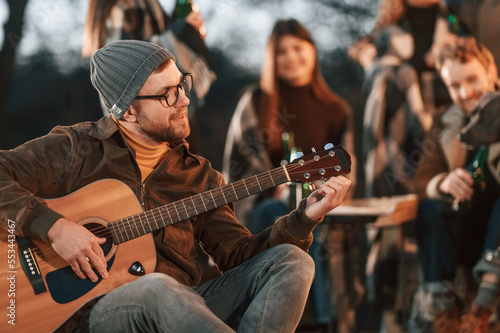Man in hat is sitting and playing guitar. Group of people is spending time together on the backyard at evening time