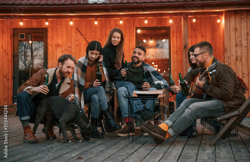 Talking, playing guitar, having fun. Group of people is together near beautiful wooden building at evening time