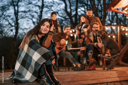 Young woman with a blanket is sitting. Group of people is spending time together on the backyard at evening time