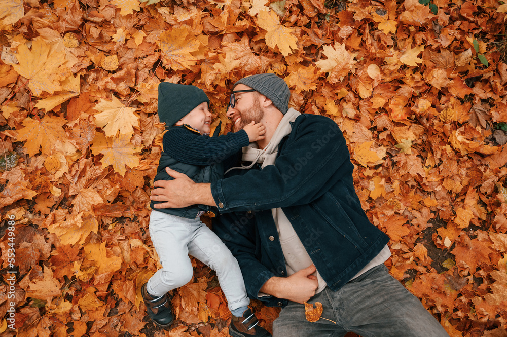 Laying down on the ground that covered with fallen leaves. Father and young son is together outdoors at daytime Laying down on the ground that covered with fallen leaves. Father and young son is together outdoors at daytime