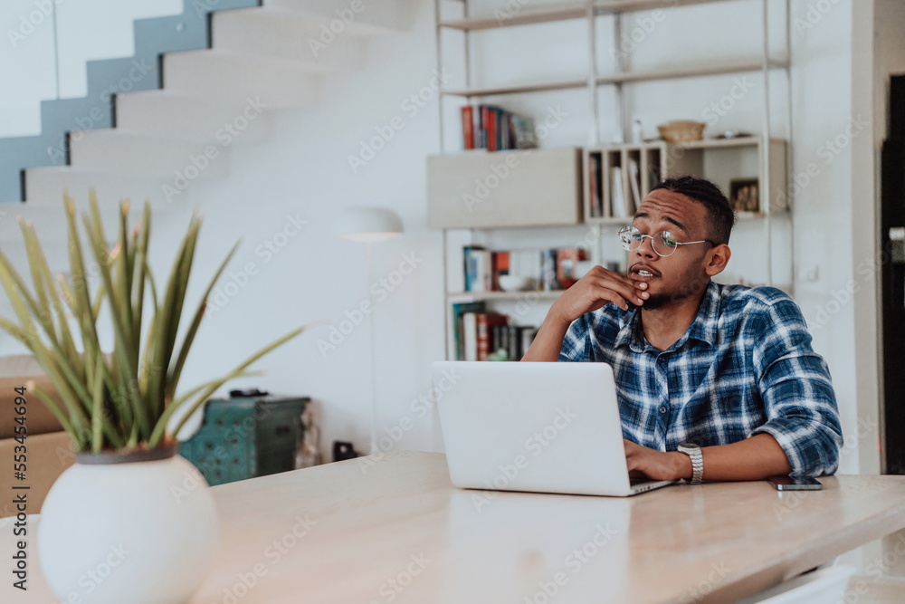 African American man in glasses sitting at a table in a modern living room, using a laptop for business video chat, conversation with friends and entertainment African American man in glasses sitting at a table in a modern living room, using a laptop for business video chat, conversation with friends and entertainment