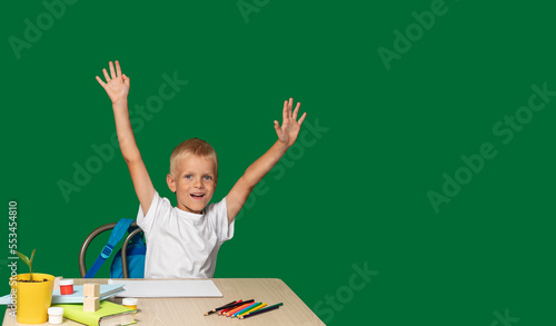 Joyful boy is sitting at table with his hands raised. There are pencils, book, cubes, paints on table. Training, upbringing, education, success. Selective focus.