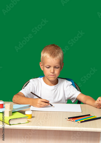 Boy looks attentively at piece of paper with pencil in his hand. There are pencils, book, cubes, paints on table. Free time, learning to draw, hobbies. Selective focus.