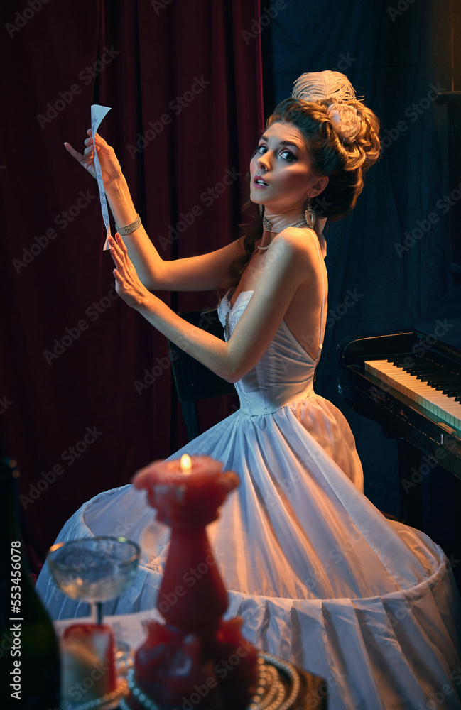 Fototapeta premium Portrait of beautiful young woman in image of medieval person, musician in tender white dress sitting at piano with new music composition. Comparison of eras, beauty, history, art
