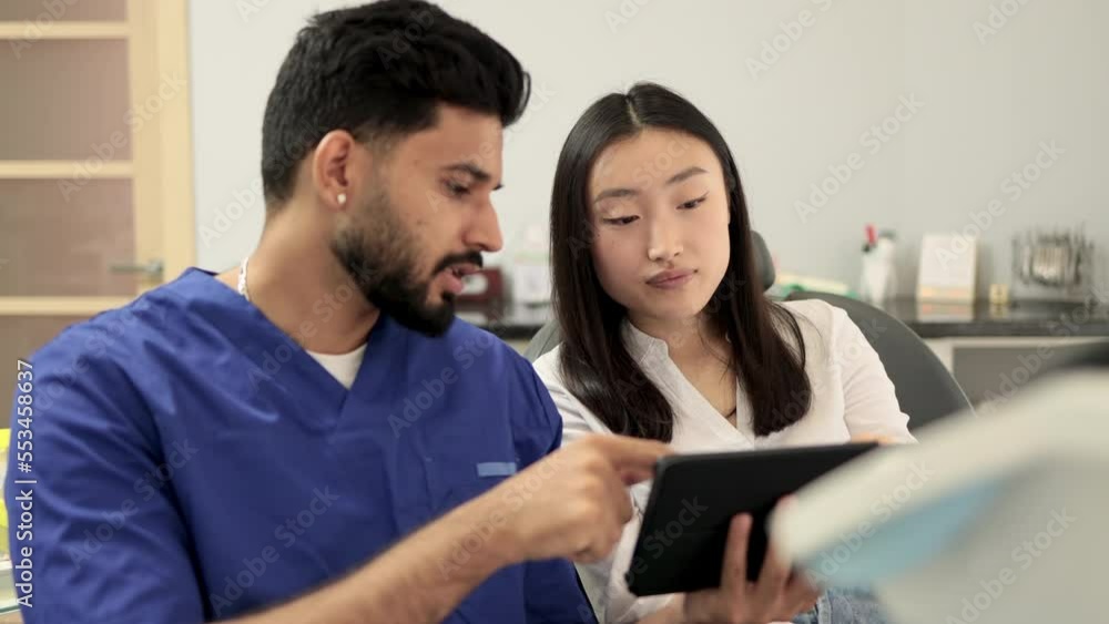 Medicine, dentistry and oral care concept. Confident bearded young man dentist showing tablet pc computer to worried female patient, asian female at modern dental clinic. Medicine, dentistry and oral care concept. Confident bearded young man dentist showing tablet pc computer to worried female patient, asian female at modern dental clinic.