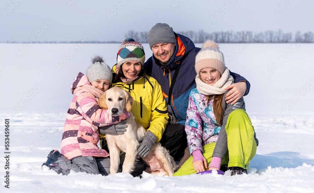 Happy family with golden retriever dog sitting on snow in winter time and smiling. Mother, father and daughters with doggy pet outdoors in Christmas season