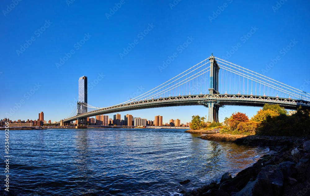 Naklejka premium Panoramic view from Brooklyn of iconic Manhattan Bridge over blue waters leading to New York City with blue sky