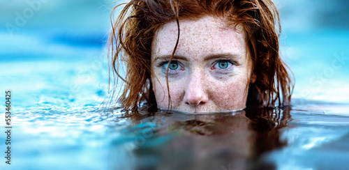 portrait of sexy, young red haired woman with freckles and red wet hair, in turquoise spa pool water, head half under water