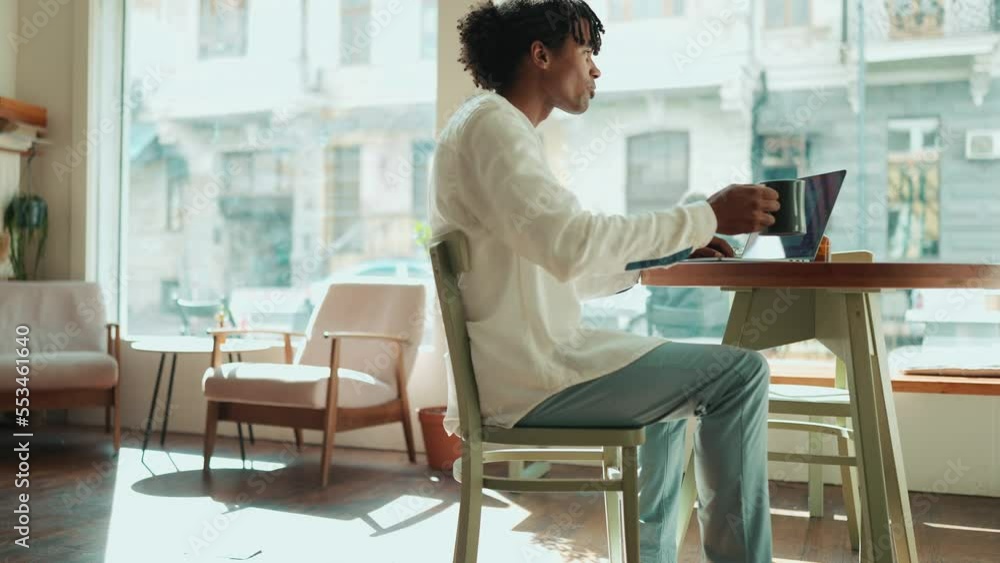 Happy African man working on laptop and drinking coffee near window in the cafe Happy African man working on laptop and drinking coffee near window in the cafe