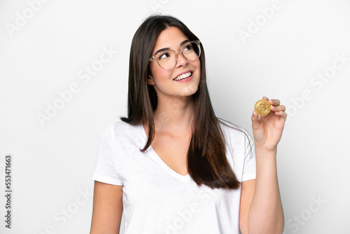 Young Brazilian woman holding a Bitcoin isolated on white background looking up while smiling