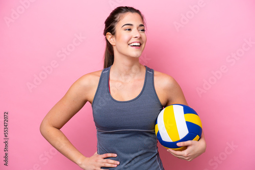 Young Brazilian woman playing volleyball isolated on pink background posing with arms at hip and smiling