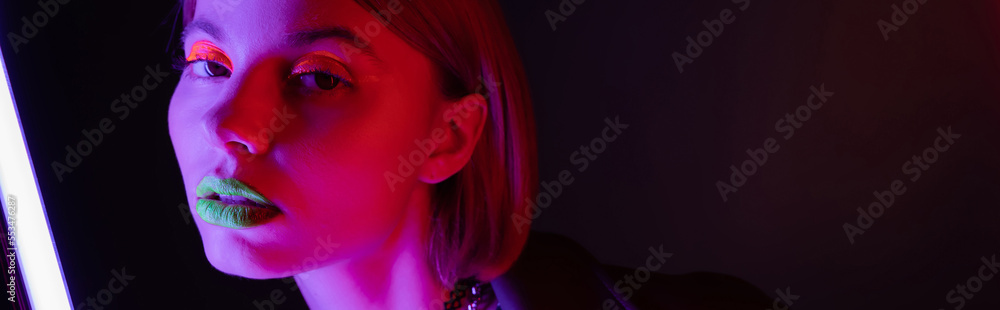 Fototapeta premium portrait of young woman with neon makeup posing in purple light of neon lamp on black background, banner.