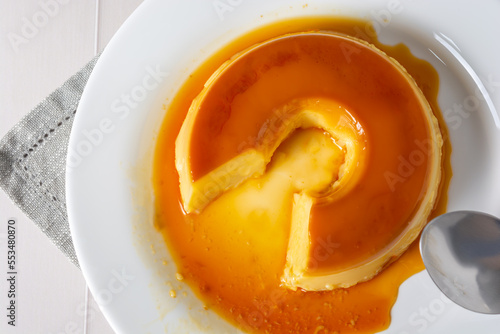 pudim de leite, also known as flan or milk pudding, isolated on white table with grey cloth. bitten slice with a metallic spoon concept of traditional brazilian dessert photo