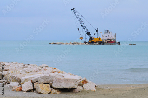 mezzo navale con gru che sistema la scogliera, vista dalla spiaggia. lavori sul molo e sulla banchina del porto con mezzi meccanici. draga photo
