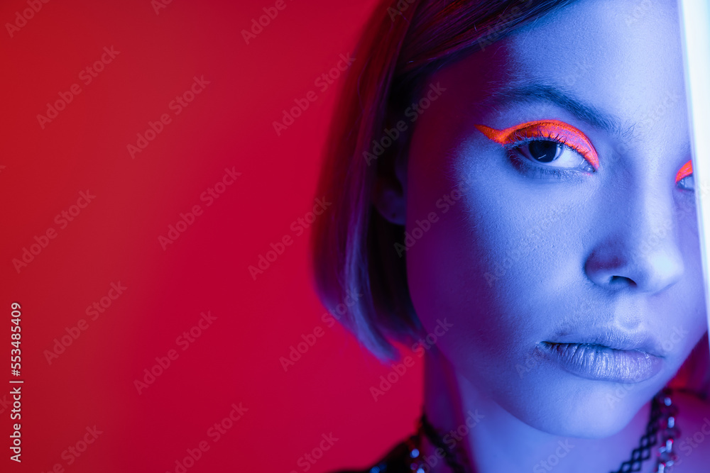 cropped view of woman with neon makeup looking at camera in blue light on carmine red background.