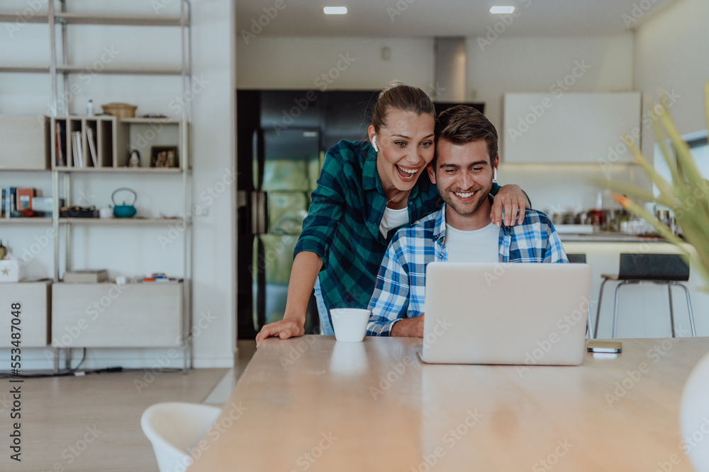 A young married couple is talking to parents, family and friends on a video call via a laptop while sitting in the living room of their modern house A young married couple is talking to parents, family and friends on a video call via a laptop while sitting in the living room of their modern house
