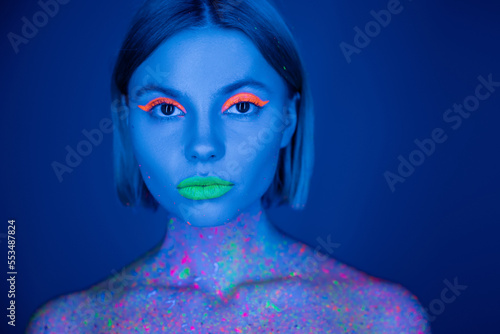 portrait of woman in vibrant neon makeup and fluorescent body paint looking at camera isolated on dark blue.