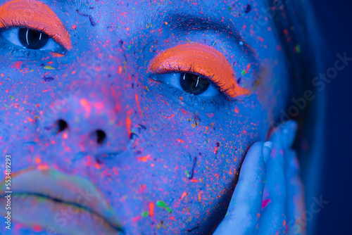 close up view of cropped woman touching face with neon makeup and bright paint splashes in blue light.