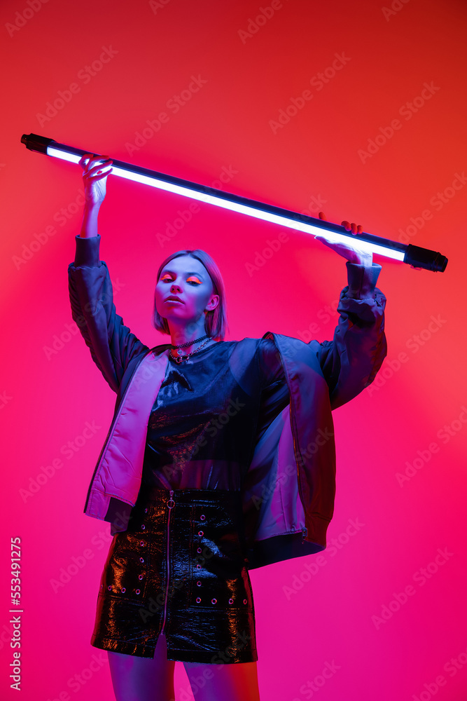 trendy woman in jacket holding glowing neon lamp above head on coral and pink background. trendy woman in jacket holding glowing neon lamp above head on coral and pink background.