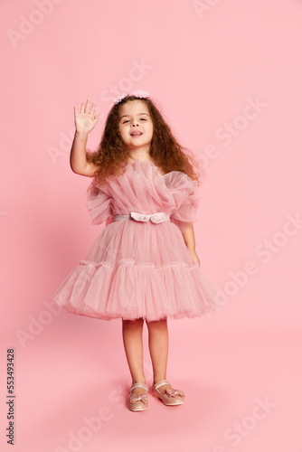 Greeting. Portrait of charming cute little girl, child with long curly hair posing isolated over pink background. Happy childhood, kids emotions, fashion and ad