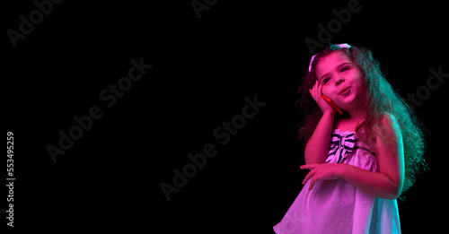Talking on phone. Stylish little girl, beginner fashion model posing isolated over dark background in neon light. Children emotions, kids fashion style, ad