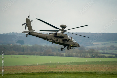 British army AH-64E Boeing Apache Attack helicopter (ZM722 ArmyAir606) moves forward from hover in to flight over a grass meadow, Wiltshire UK 