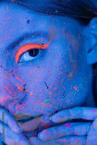 partial view of woman holding hands near face with neon makeup and bright paint splashes in blue light.