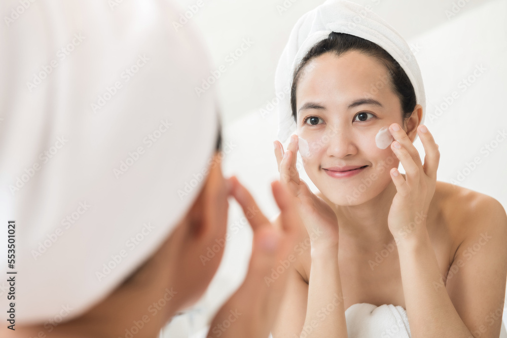 Happy young Asian woman applying face lotions while wearing a towel and touching her face in bathroom Happy young Asian woman applying face lotions while wearing a towel and touching her face in bathroom